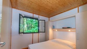Una habitación pequeña y acogedora con una cama, un ventanal con vistas a un bosque y una estantería con un libro.