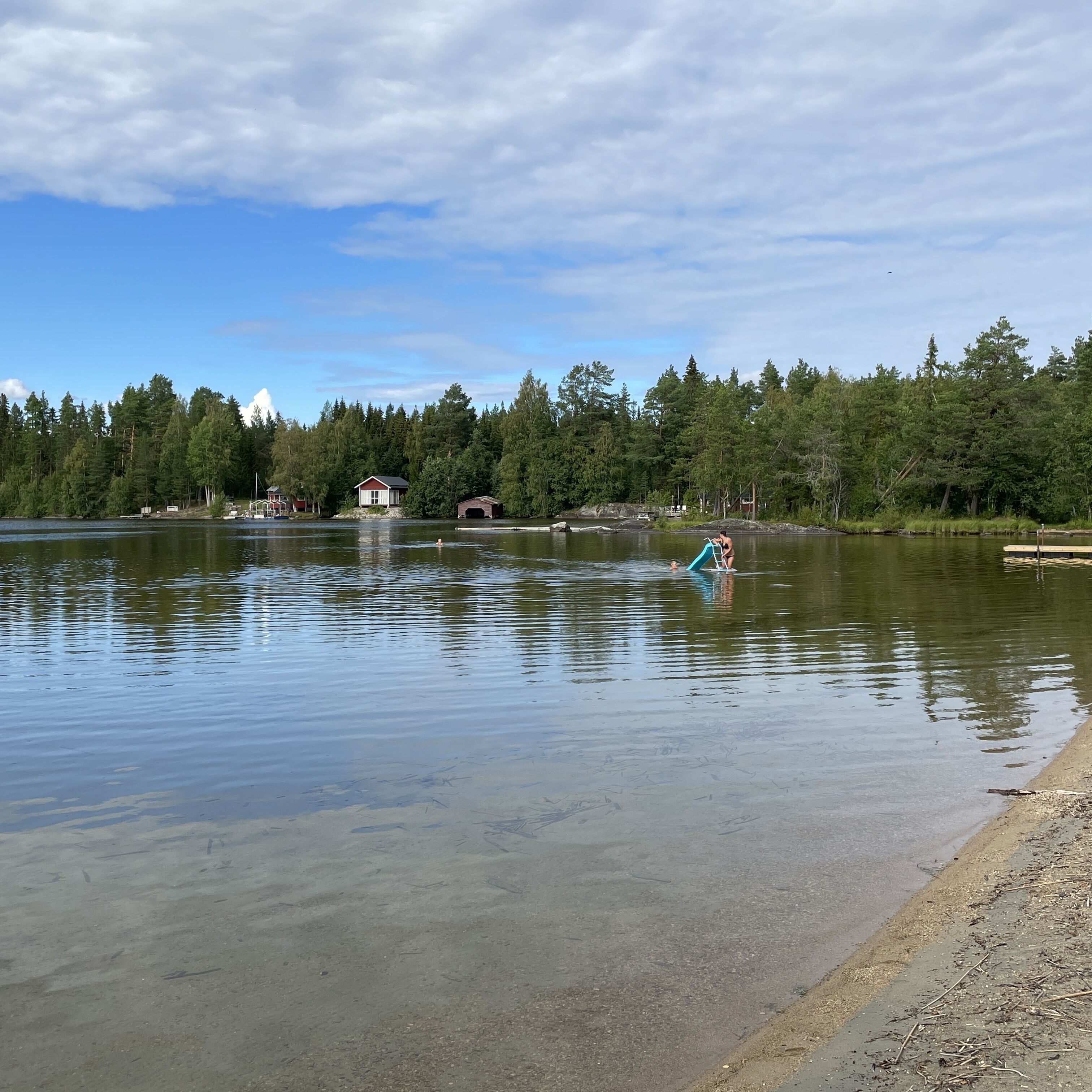 A calm lake with a wooden dock extending into the water, surrounded by dense forest and a few buildings in the background.