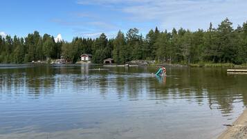 A calm lake with a wooden dock extending into the water, surrounded by dense forest and a few buildings in the background.