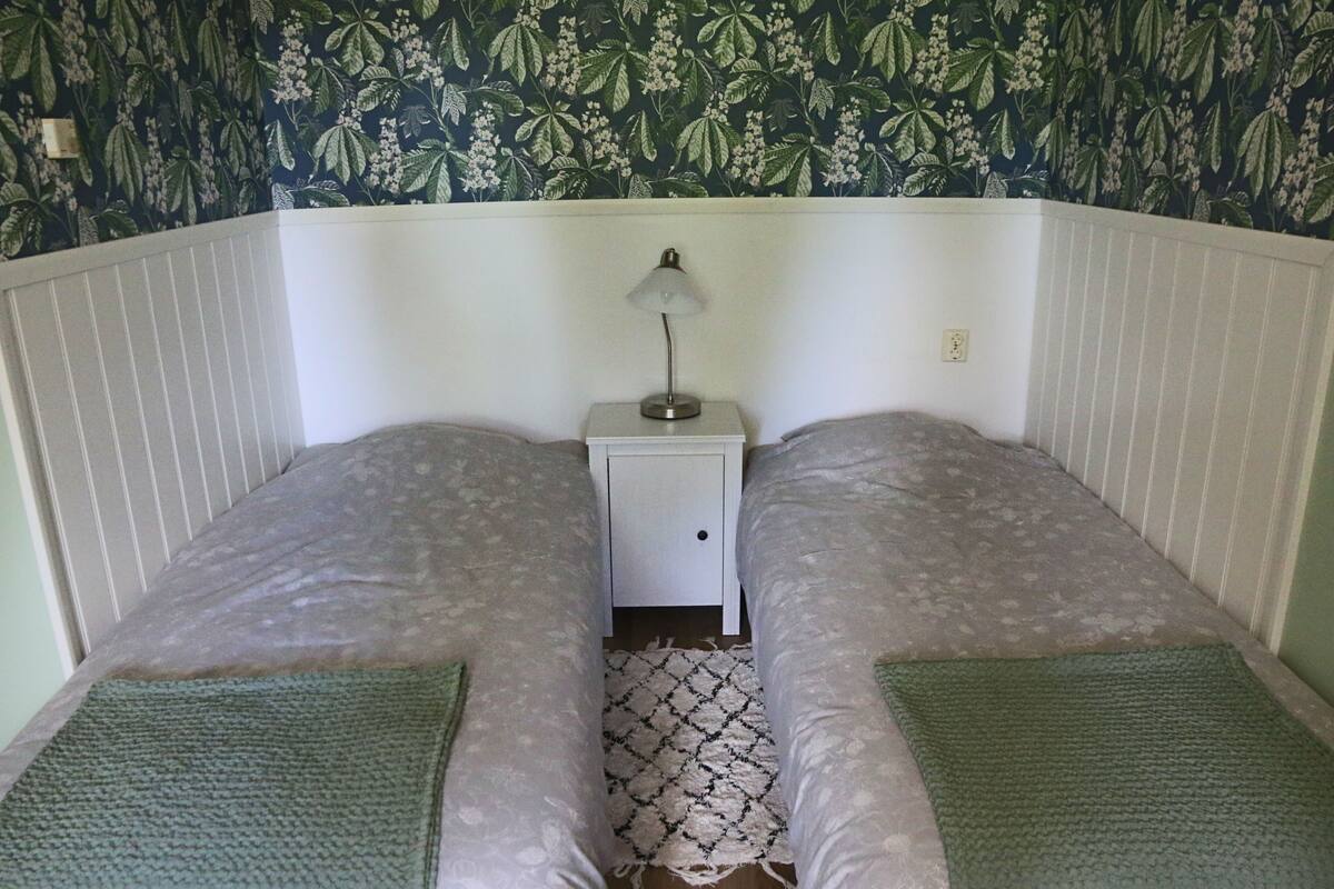 A twin room with two beds, a nightstand with a lamp, a patterned wallpaper, and a small rug.