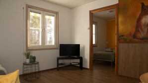 Una sala de estar con televisor sobre un mueble bajo, una mesita auxiliar con una planta, un ventanal con vistas a árboles y un cuadro en la pared.