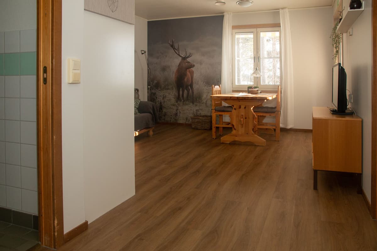A room with a wooden floor, a dining table with chairs, a sofa, a wall-mounted lamp, and a window with curtains.