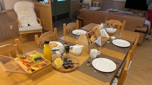 Una mesa de comedor dispuesta con platos, cubiertos, salsas y una cesta de pan en una habitación acogedora con muebles de madera y una zona de estar confortable.