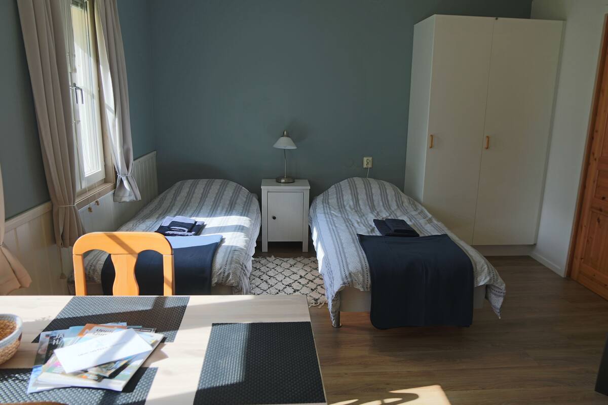A room with two single beds, a small table with a chair, a lamp, and a wardrobe.