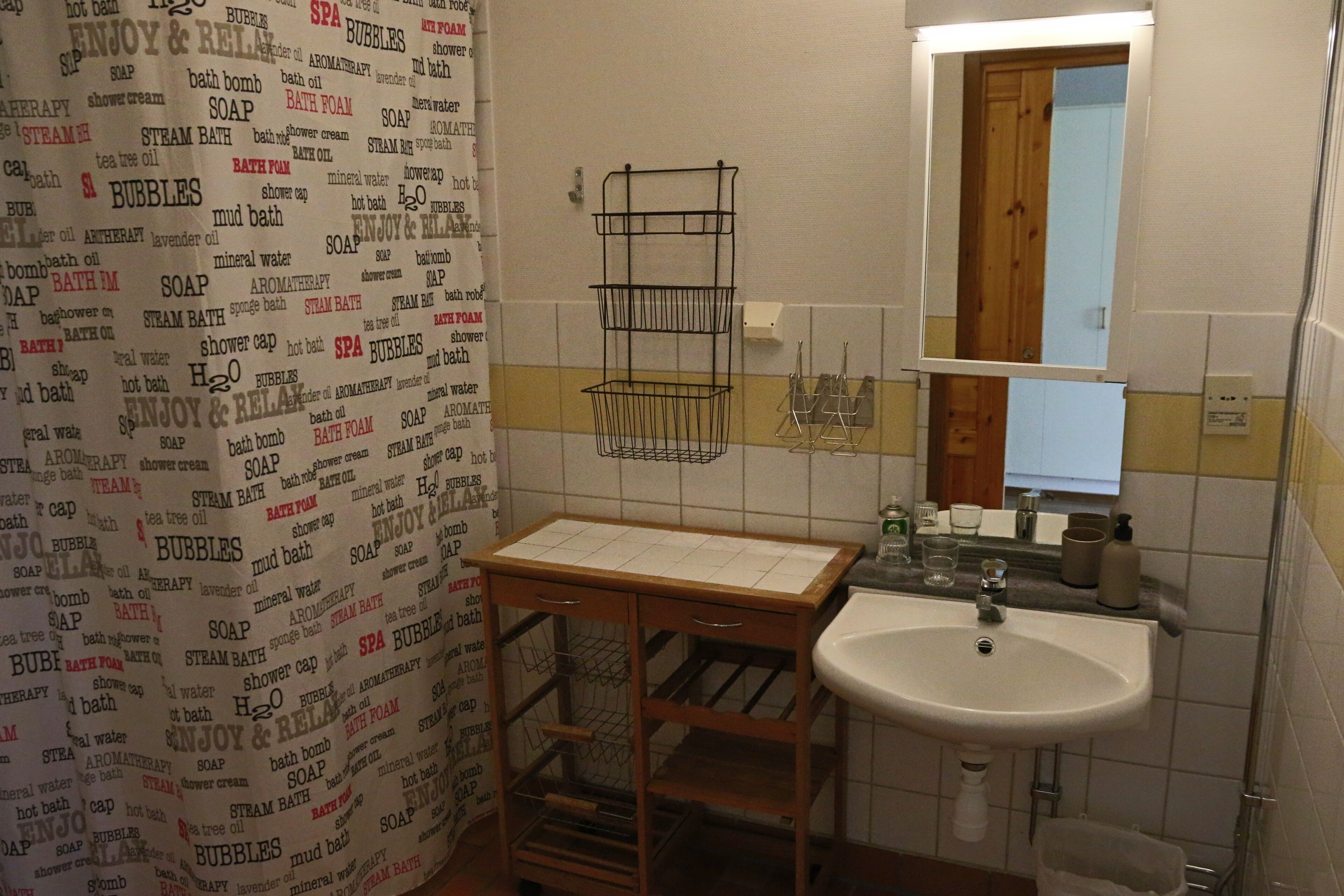 A bathroom with a sink, mirror, and towel rack. There is a shower curtain with text and a wooden shelf with a white sink.