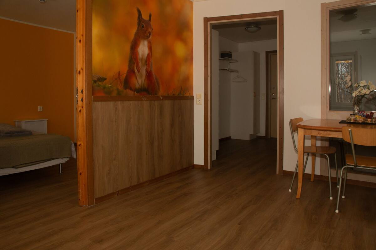 A room with a wooden floor, a dining table with chairs, a bed with a green bedspread, and a painting of a squirrel on the wall.