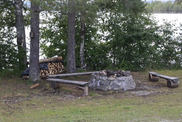 Un entorno rústico al aire libre con un hogar de piedra, bancos de madera y leña apilada.