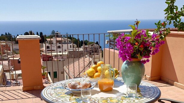 Una mesa redonda con mantel floral, un florero con flores, un cuenco de limones, una jarra de jugo de naranja y un plato con pasteles en un balcón con vista al mar.