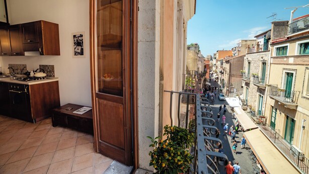 Uma rua estreita com edifícios em ambos os lados, uma varanda com grade de metal e vista para a rua abaixo.