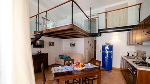 Uma sala de jantar com mesa de madeira, cadeiras e uma tigela de frutas. Há uma geladeira azul e uma cozinha com armários de madeira e fogão. Uma escada com corrimão de vidro leva a um andar superior.