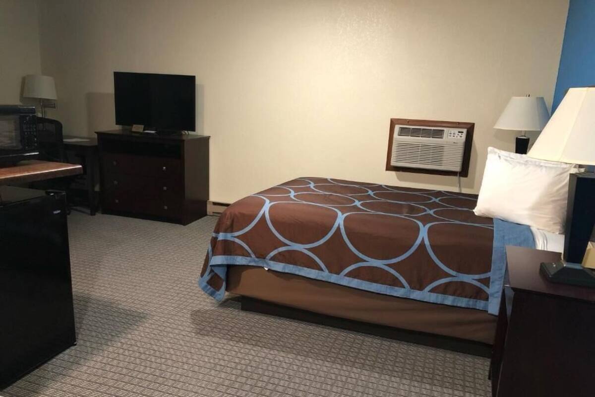 A hotel room with a bed, a desk with a computer, a television, a lamp, and a wall-mounted air conditioner.