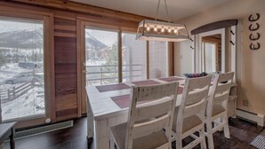 Ein Essbereich mit Holztisch und Stühlen, großen Fenstern mit Jalousien und einer Pendelleuchte.