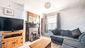 A living room with a television on a wooden stand, a sofa, a coffee table, and a fireplace. - Cosy Welsh Cottage in St Davids Centre with Garden (St Davids)