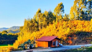 A small wooden cabin with a red roof by a calm river, surrounded by autumn-colored trees and a clear sky. - Cabana Brisa Serrana - Urubici-SC (Urubici)