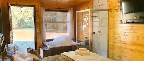 A wooden cabin bedroom with a large bed, a separate sleeping area, a glass shower, and a view of a natural landscape.