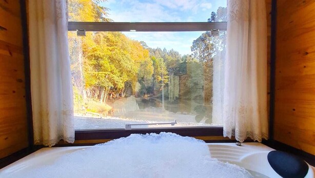 A wooden cabin room with a large bathtub filled with water, a window with curtains, and a view of a forest and lake. - Cabana Brisa Serrana - Urubici-SC (Urubici)