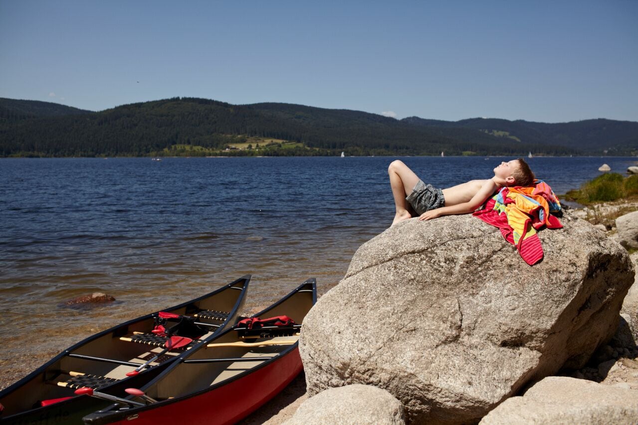 A person relaxing on a rock by a lake with canoes nearby.