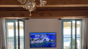 A room with a wooden ceiling, a chandelier, a large TV on the wall, a table with two bottles, and a bench. - Antoine (Bonifacio)