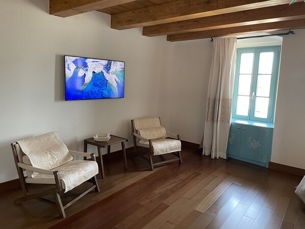 Une pièce avec un plancher en bois, deux fauteuils rembourrés, une petite table, un tableau accroché au mur et une fenêtre avec des rideaux.