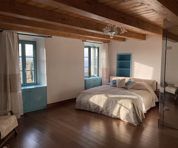 A bedroom with a wooden floor, a bed with white bedding, a blue cabinet, and a chandelier. - Antoine (Bonifacio)