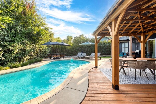 Une piscine dotée d’une terrasse en bois et de chaises longues, entourée de verdure.