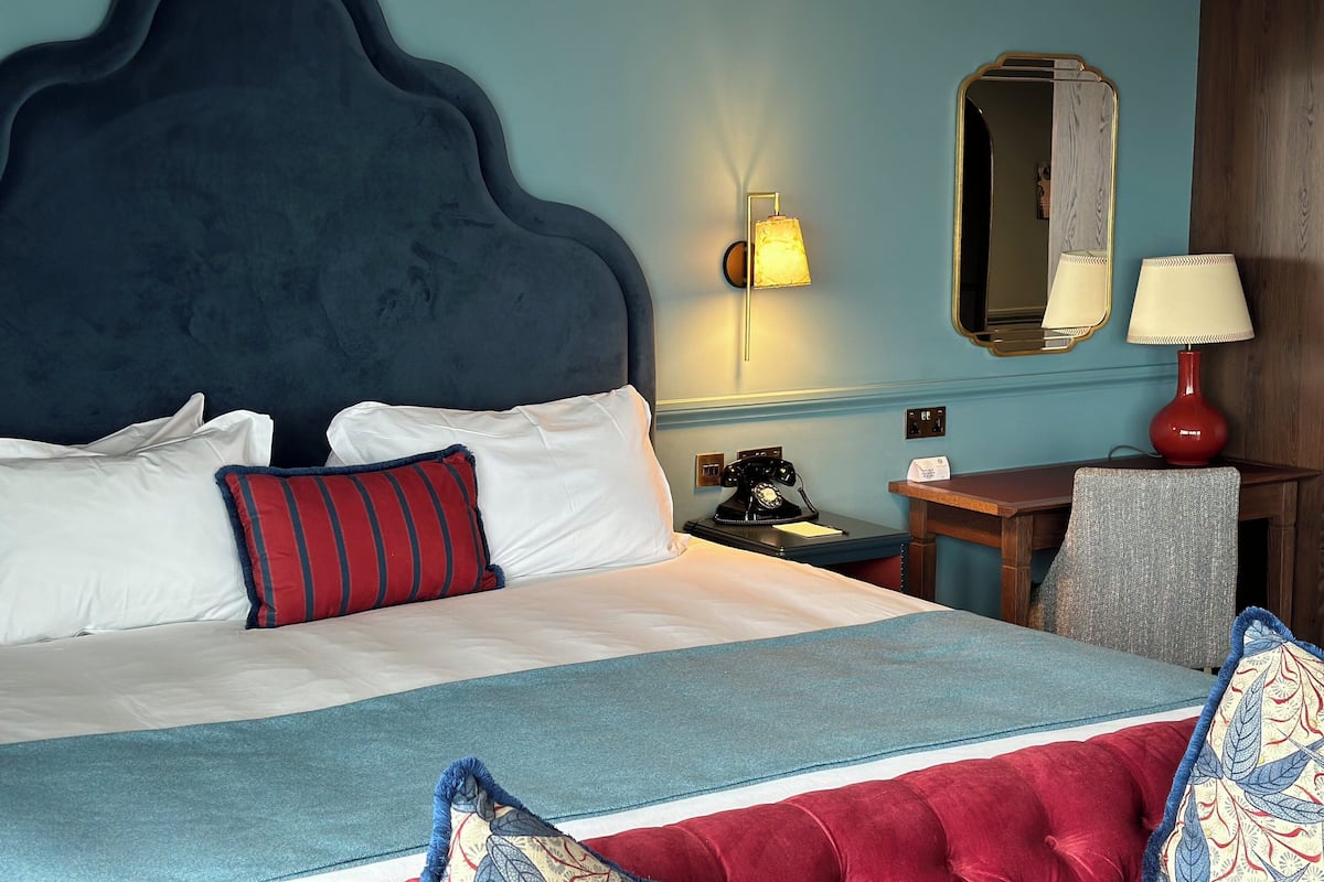 A neatly made bed with white and red pillows, a blue headboard, a bedside table with a lamp, and a mirror on the wall.