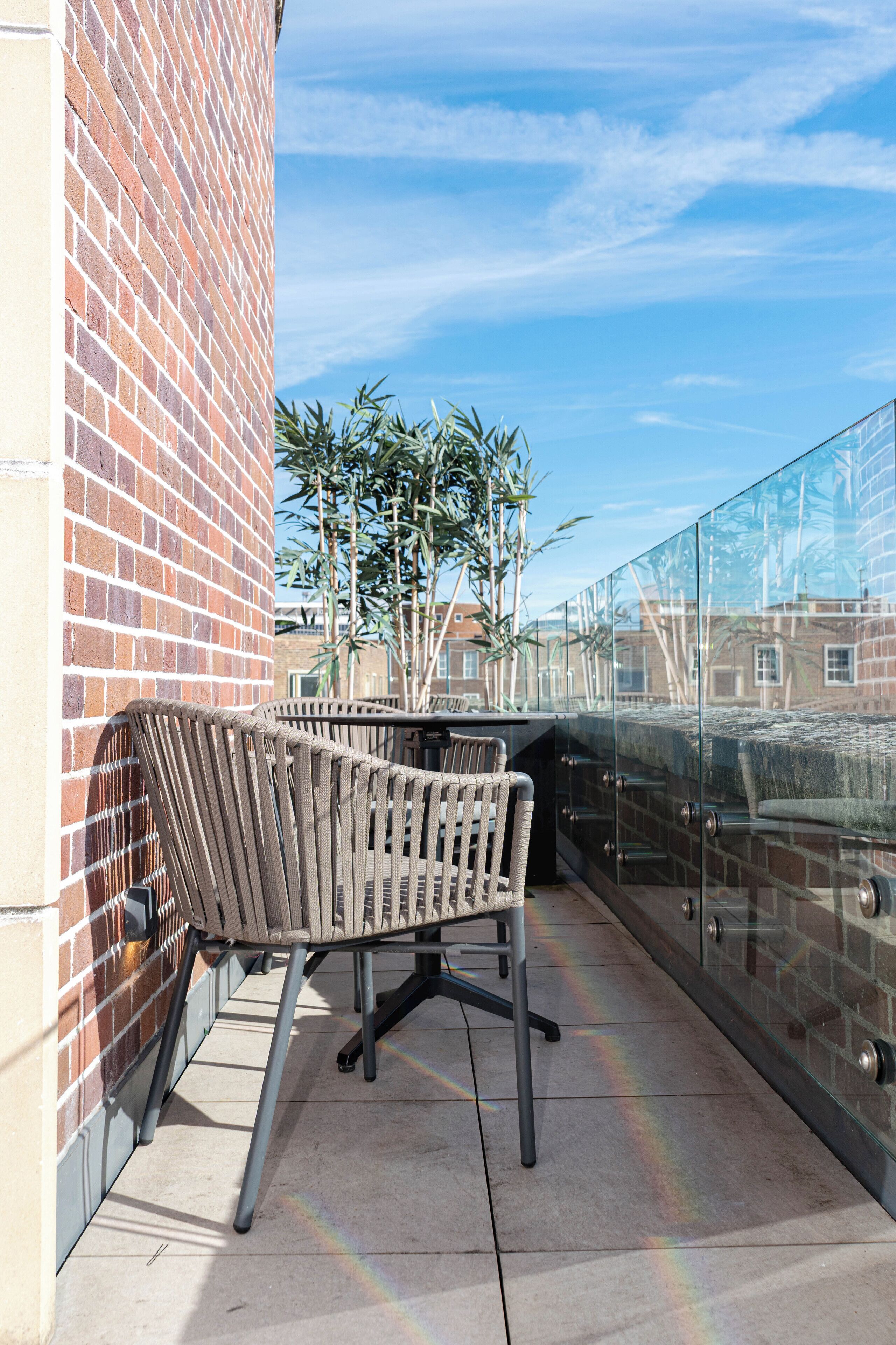 Een stoel met metalen frame op een betegeld balkon met glazen balustrade, een bakstenen muur en een bamboe plant op de achtergrond.