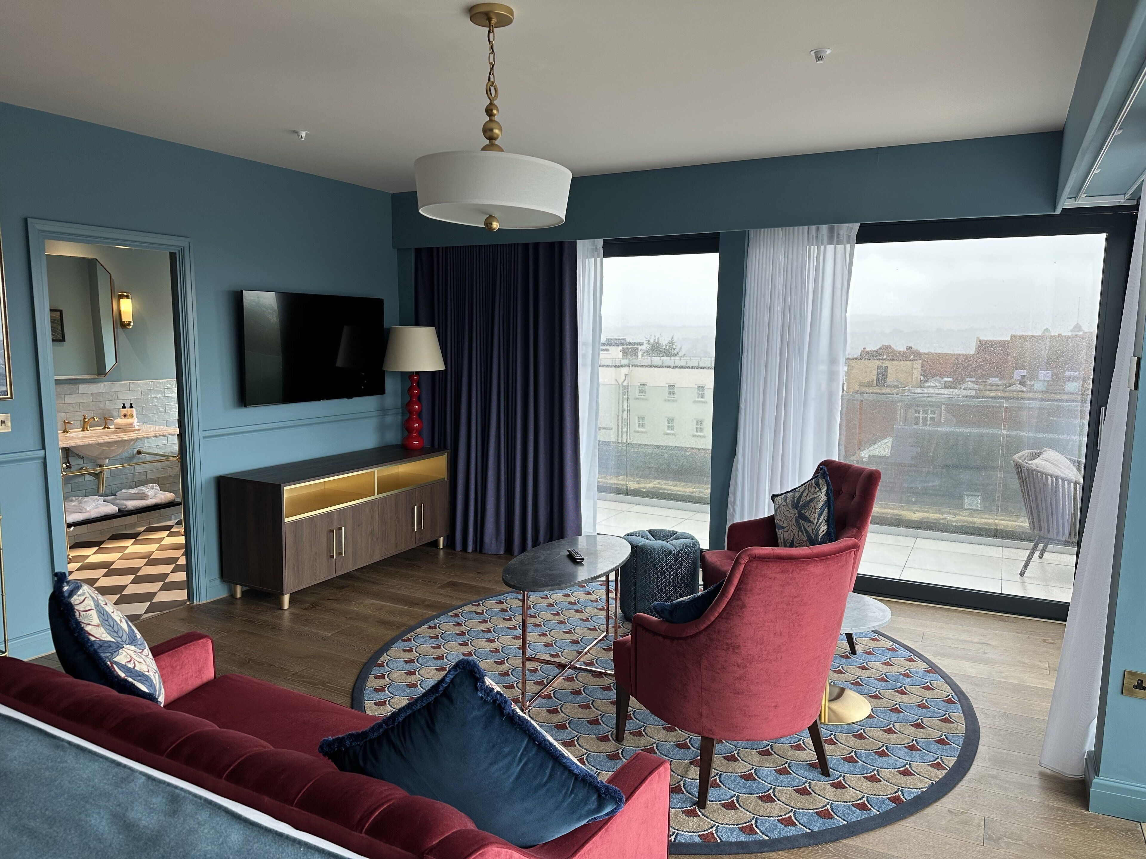 A modern living room with a red sofa, a red armchair, a round coffee table, a TV, and a large window with a city view.