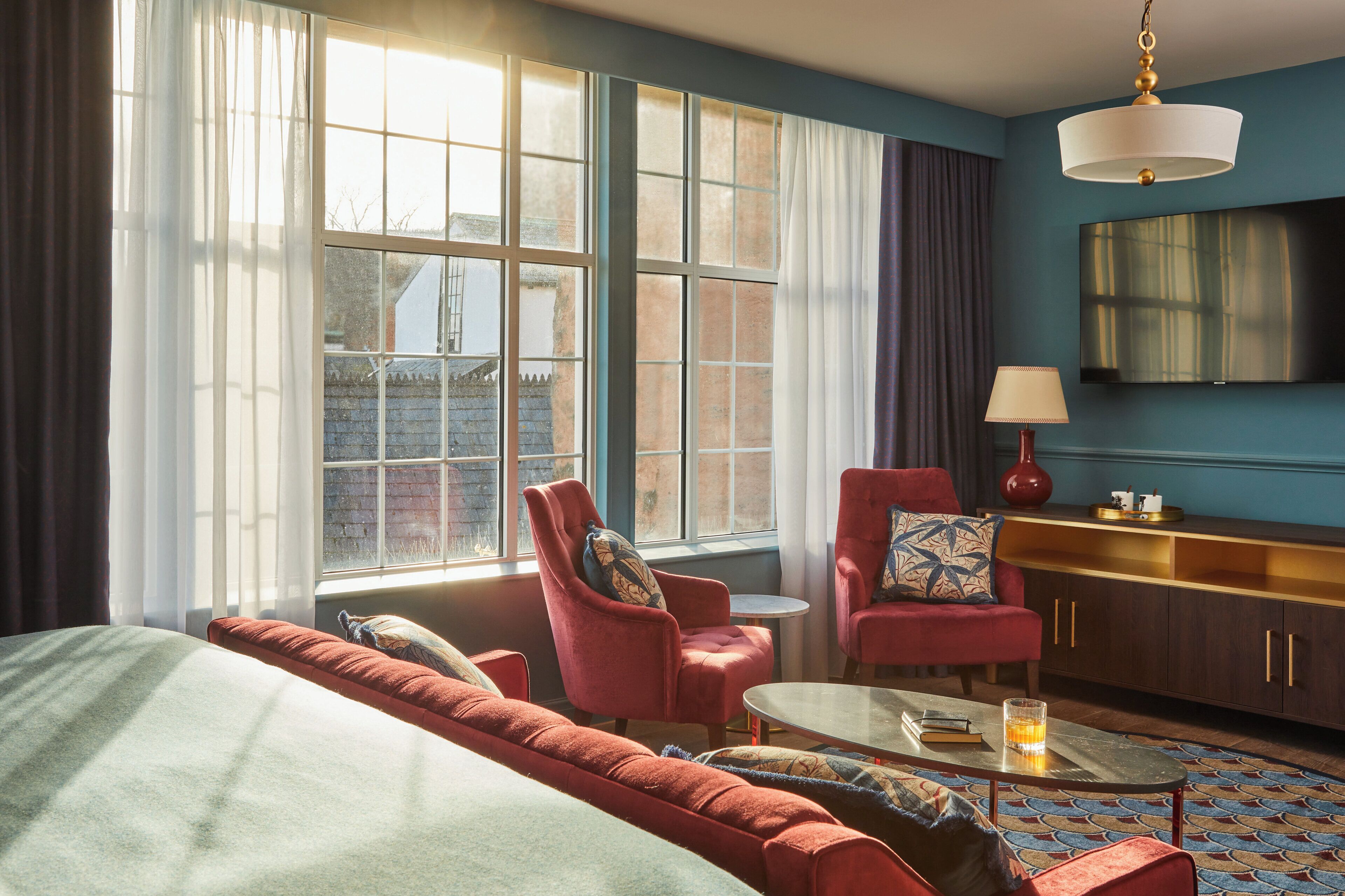 A hotel room with a large window, a sofa, a chair, a small table, and a lamp.