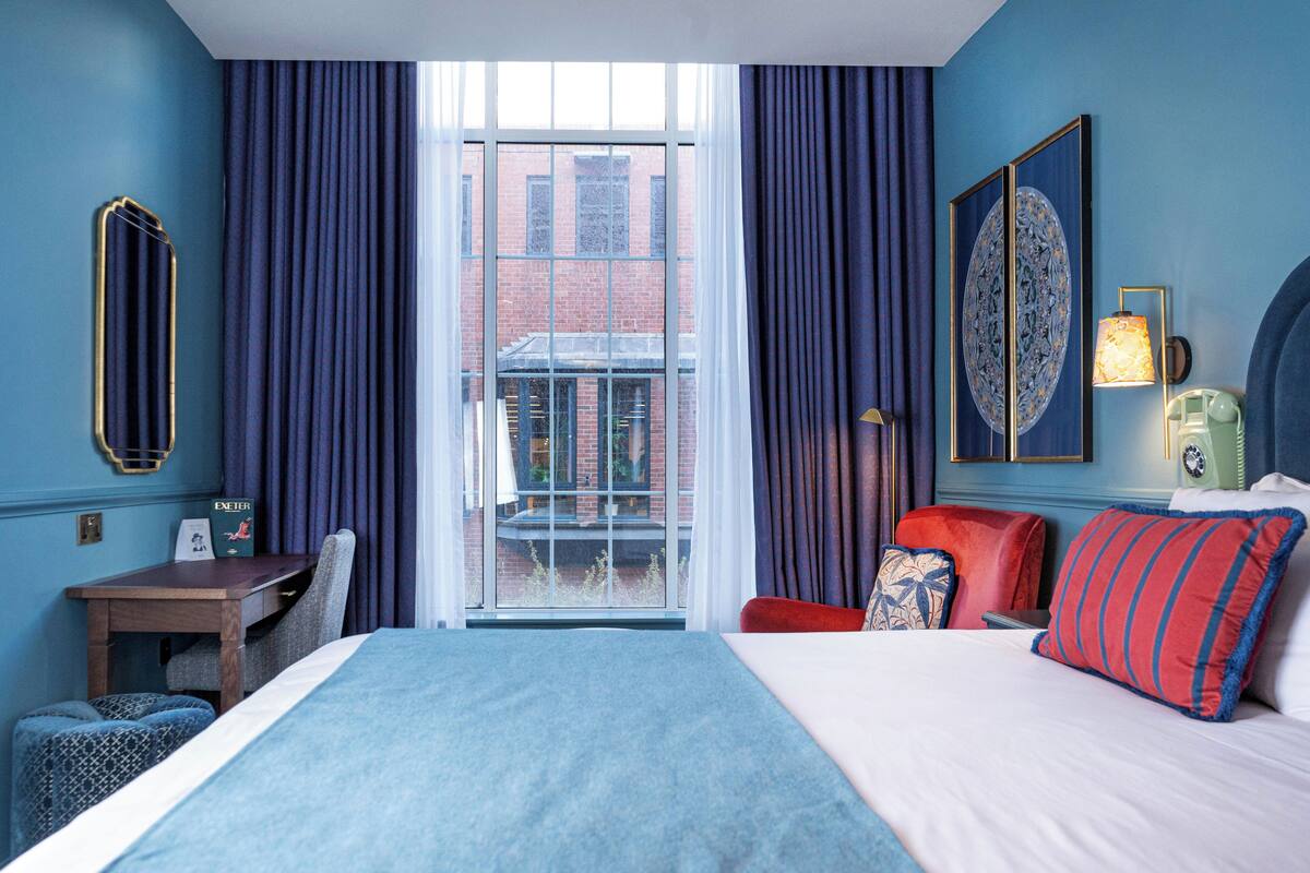 A hotel room with a bed, a desk, a chair, a red sofa, and a view of a building through a window.
