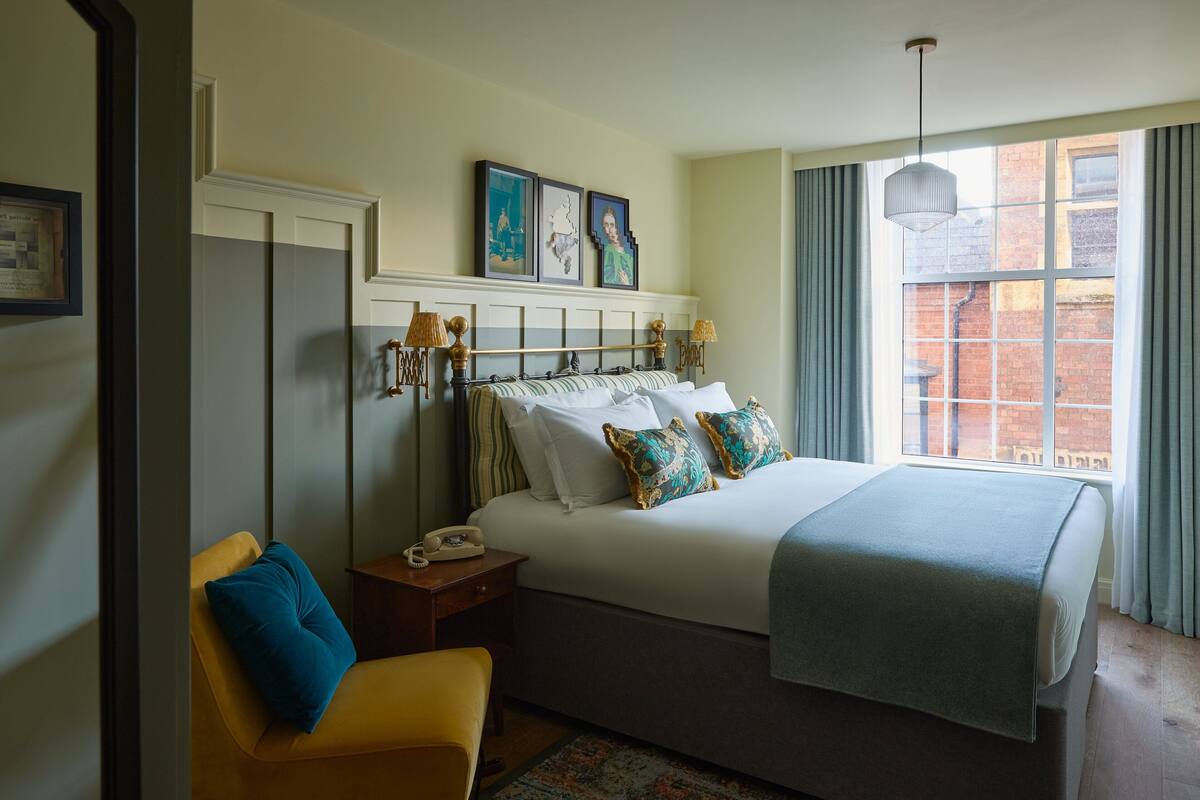 A hotel room with a large bed, a bedside table with a lamp, a chair, and a window with curtains.