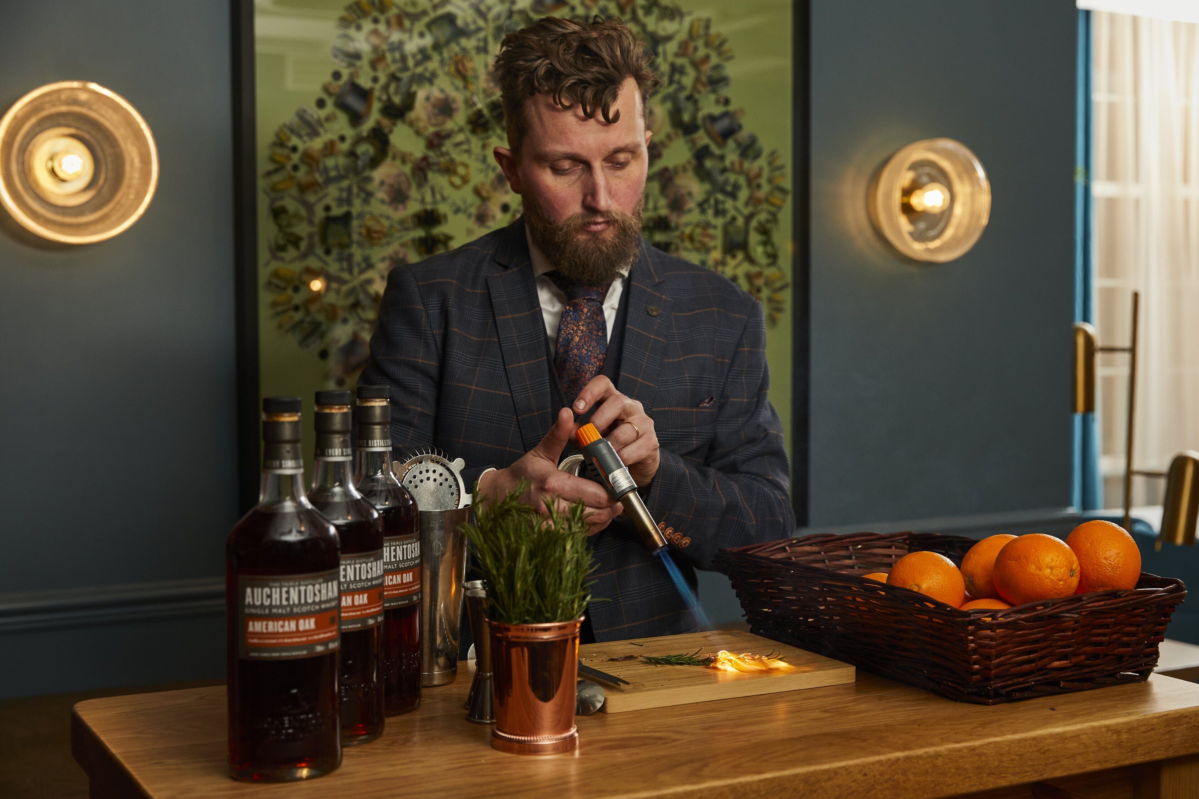 A man in a suit preparing drinks at a bar with bottles and a basket of oranges.