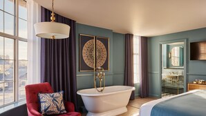 A room with a large bathtub, a red armchair with a patterned pillow, a bed with a blue bedspread, and a large mirror above the sink.