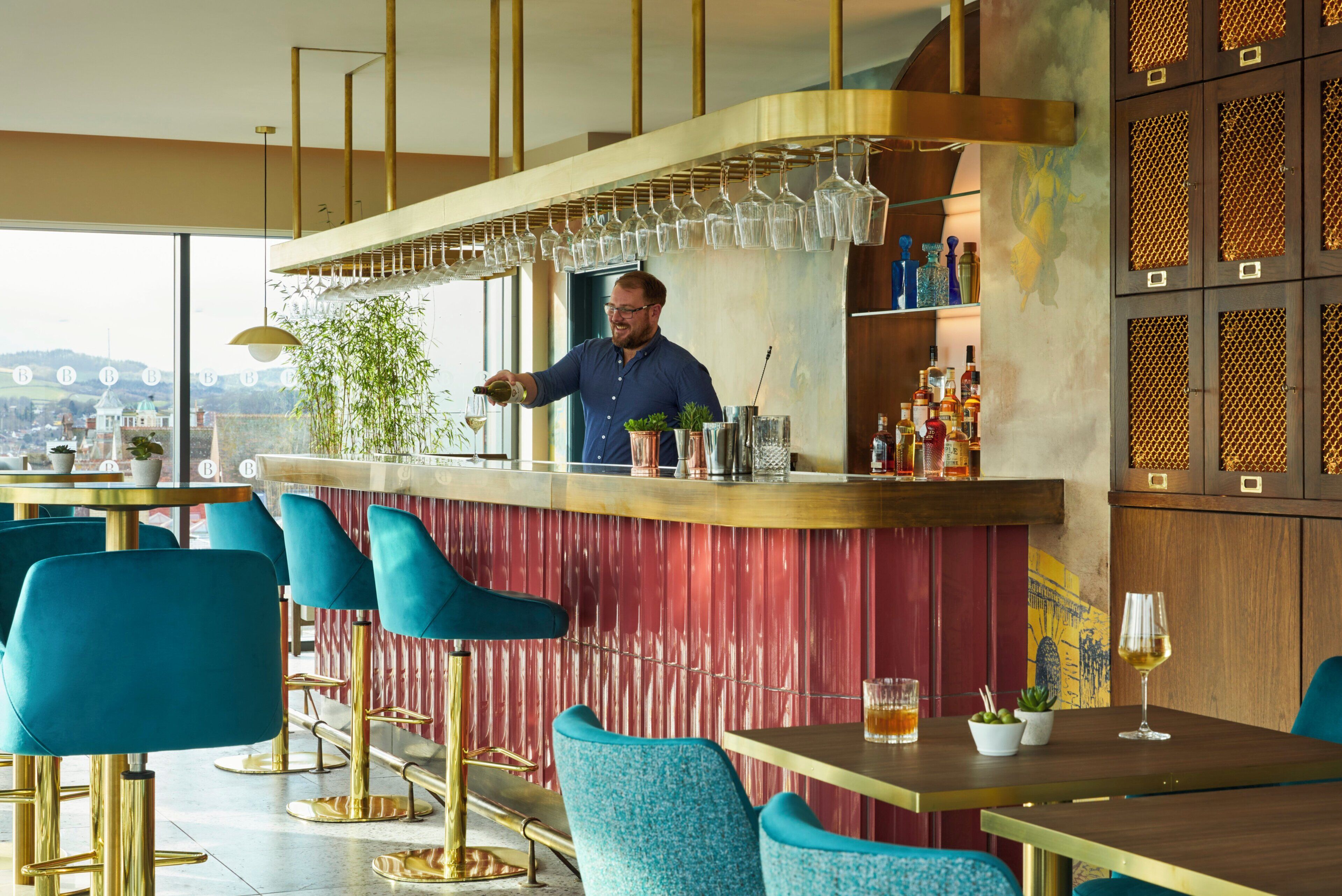 A bar area with a bartender, blue bar stools, and a view of the city.