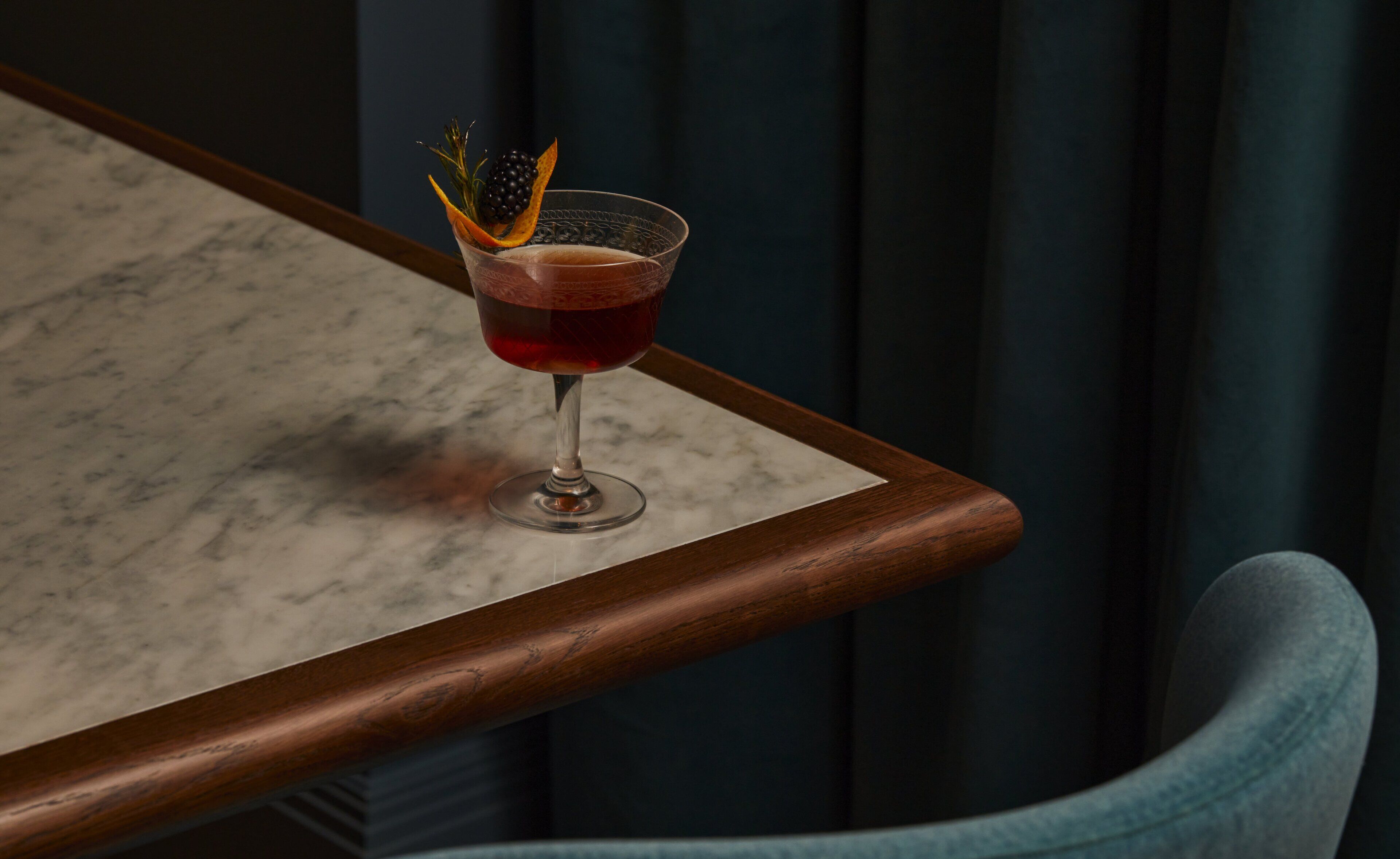 A bar setting with a glass of red drink garnished with a black berry and an orange peel, placed on a marble table with a wooden edge.