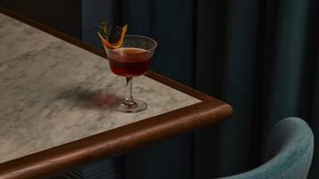 A bar setting with a glass of red drink garnished with a black berry and an orange peel, placed on a marble table with a wooden edge.