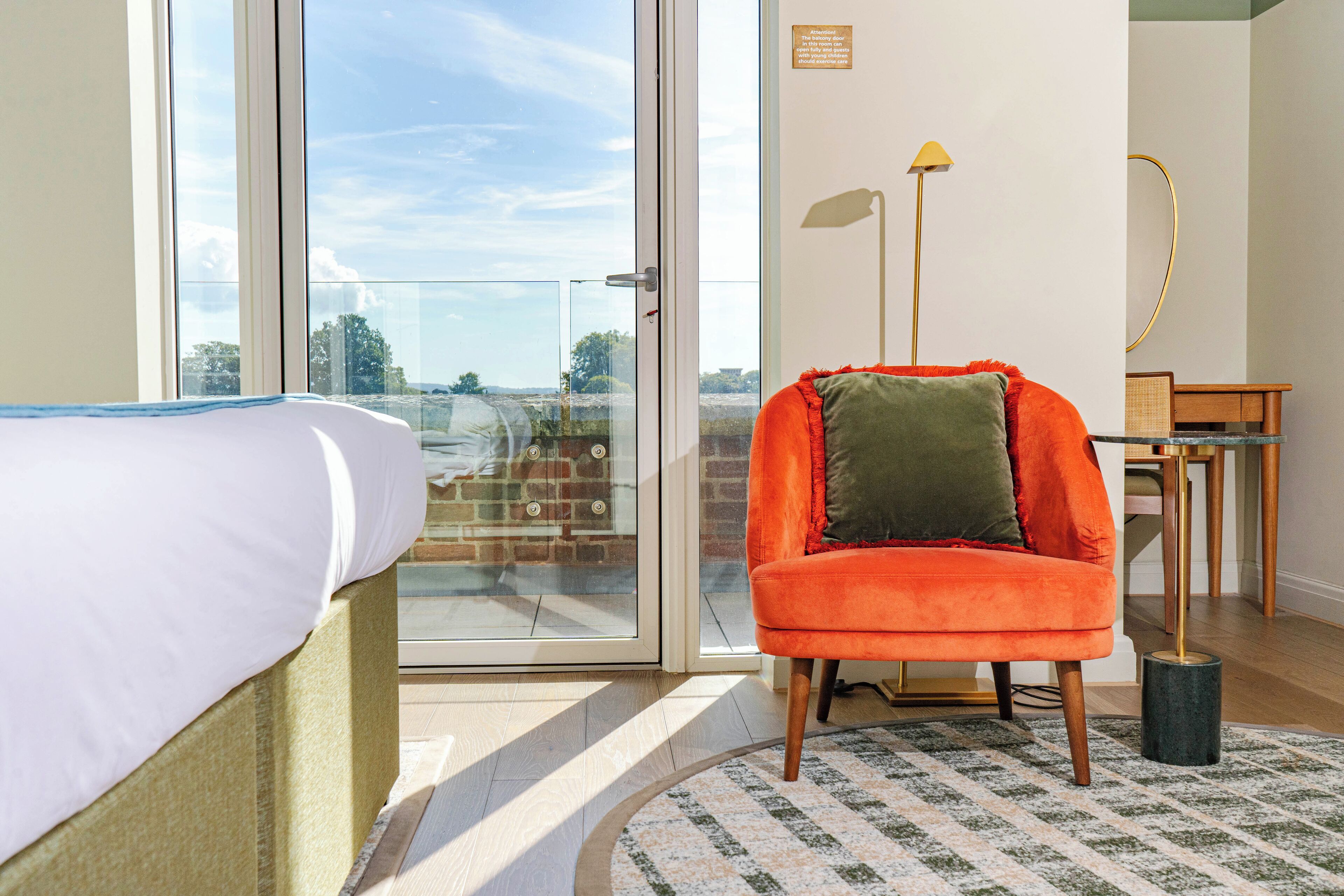 A hotel room with a bed, a bright orange chair, a small table, and a view through a glass door.