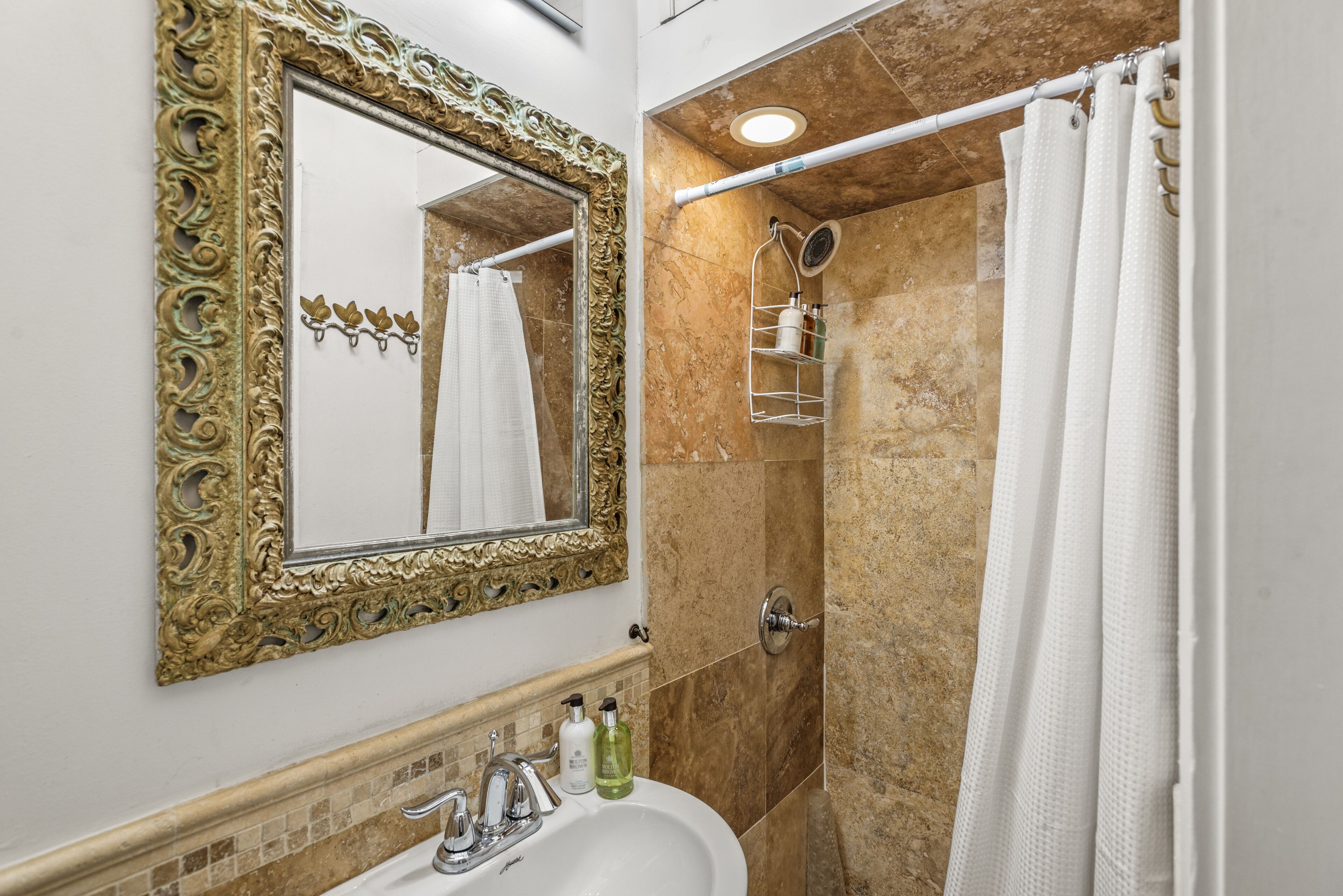 Un baño con un espejo grande y ornamentado, un lavabo con dos grifos, una ducha con puerta de vidrio y un perchero para toallas.