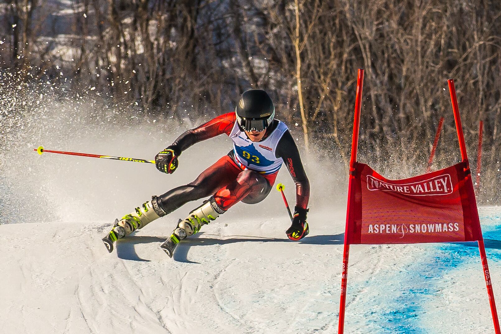 Un esquiador en movimiento, con un dorsal, esquía por una ladera nevada con una bandera roja al fondo.