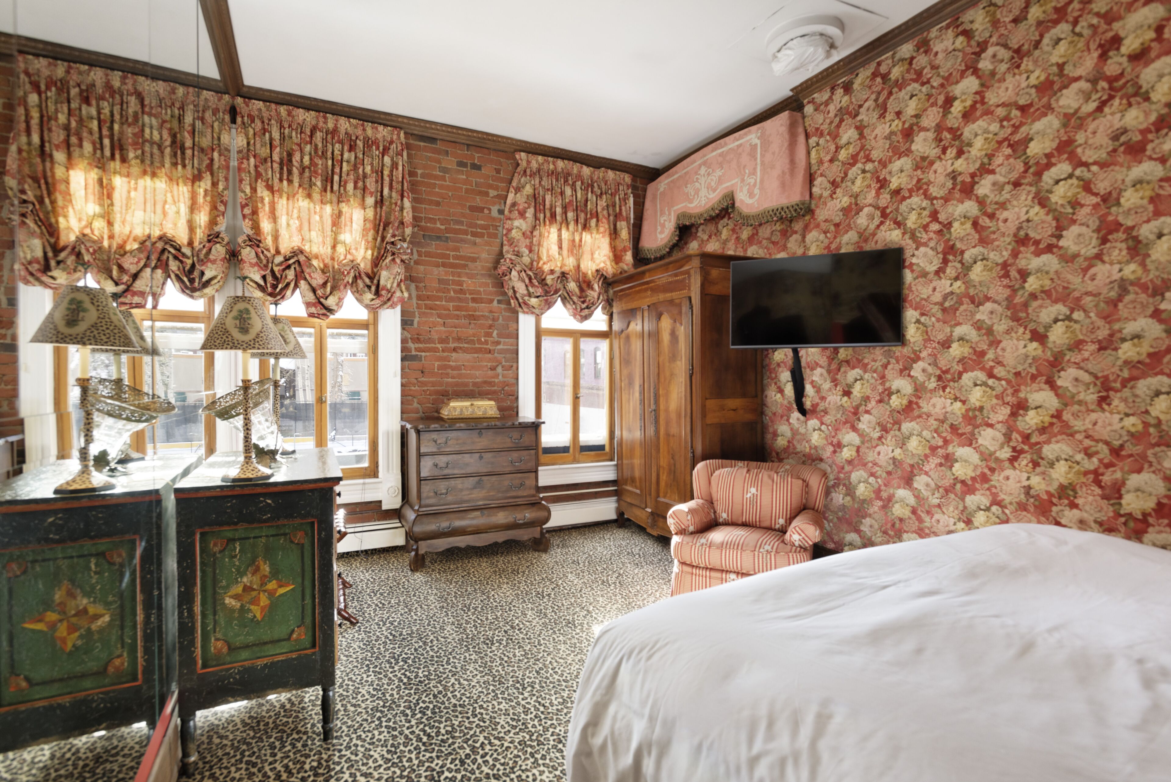 Une chambre à coucher comprenant un lit, une commode, une chaise, un téléviseur et une fenêtre avec des rideaux.