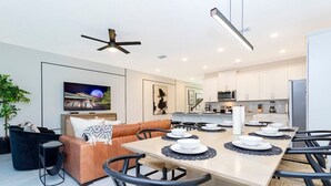 Eine moderne Küche mit weißen Schränken, ein Essbereich mit einem langen Tisch und ein Wohnzimmer mit einer Couch und einem Fernseher.