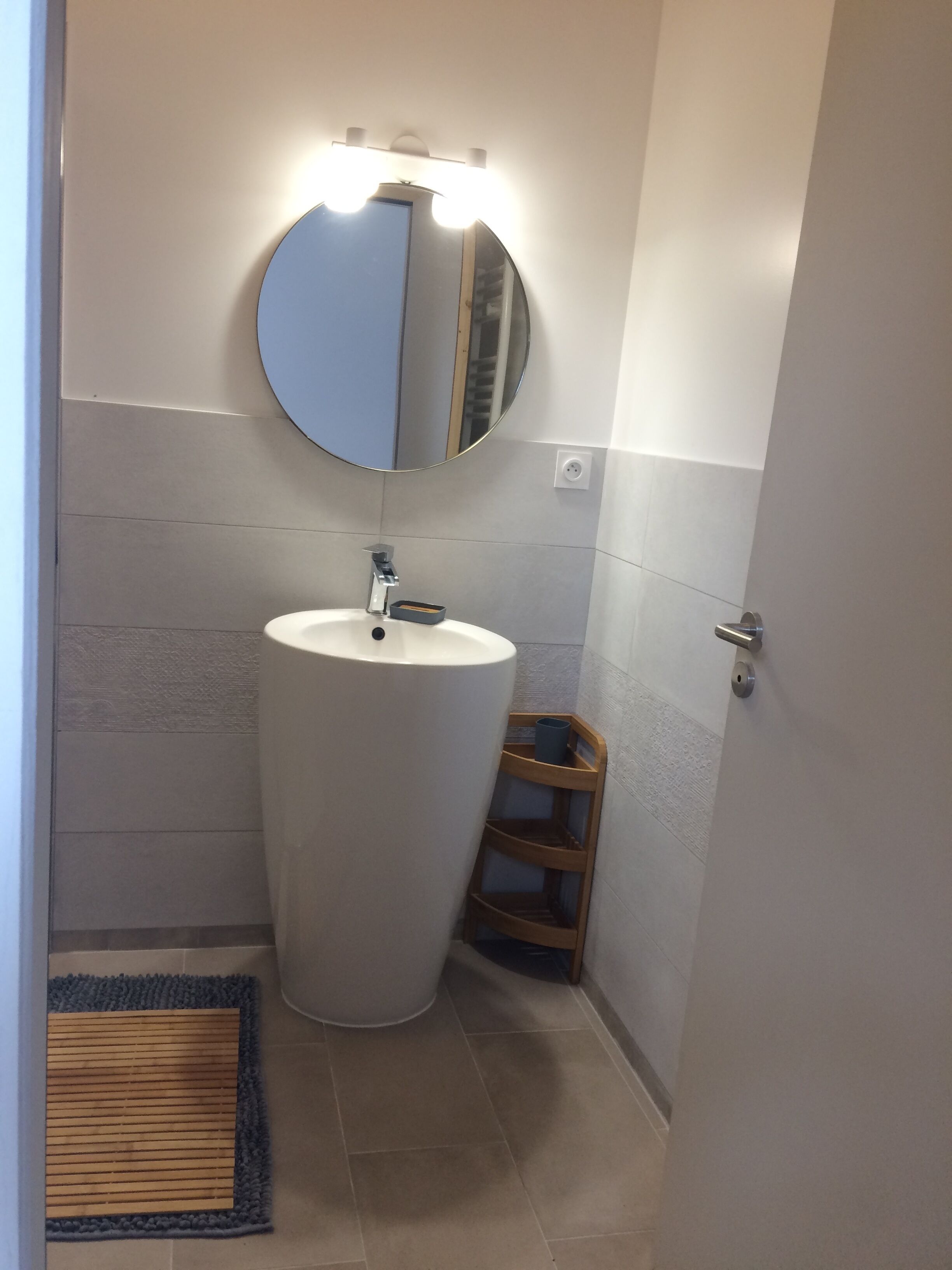 A bathroom with a large white sink, a round mirror, and a small wooden shelf.