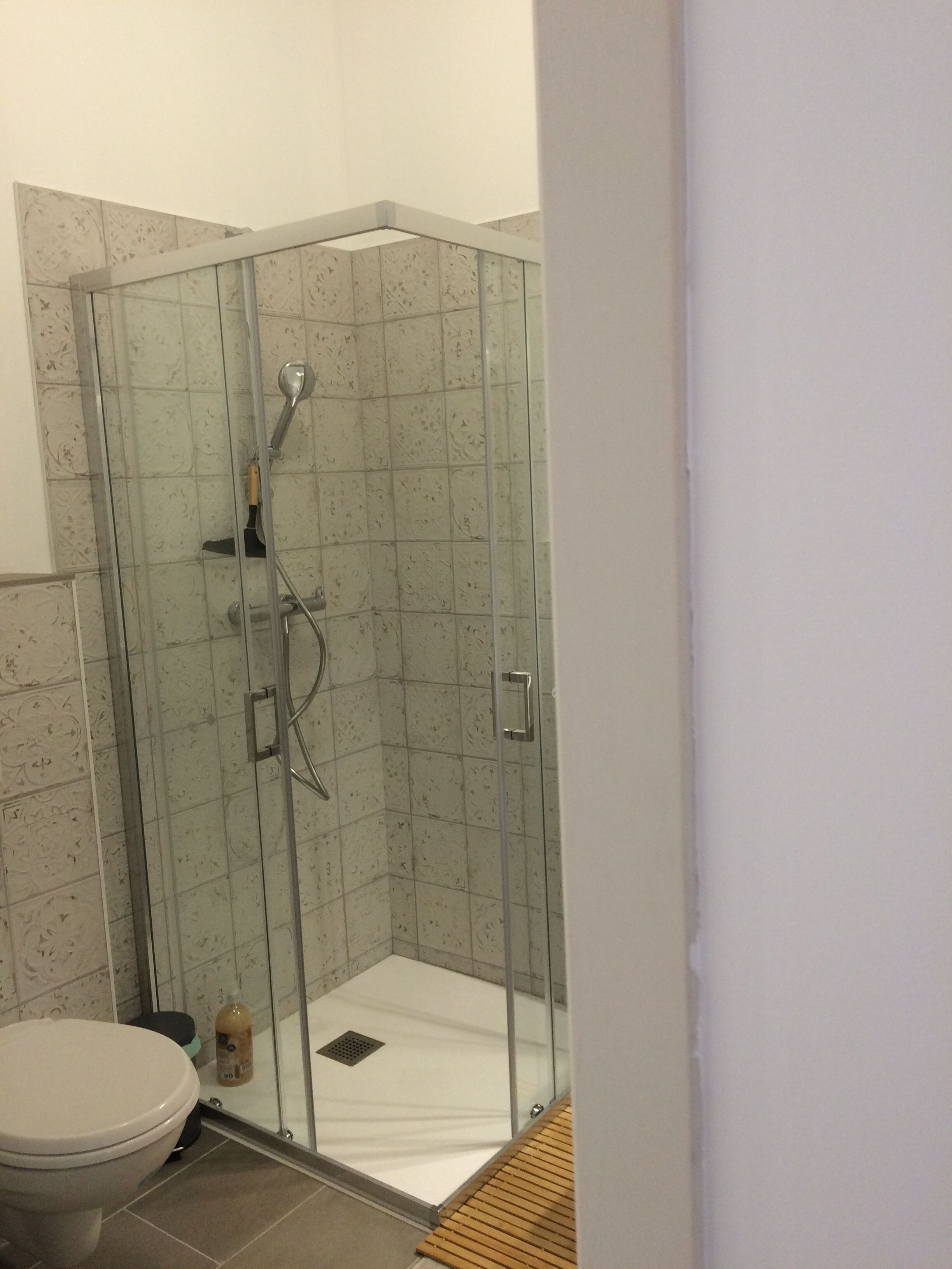 A glass-enclosed shower stall with a white floor, a toilet, and a bottle of shampoo.