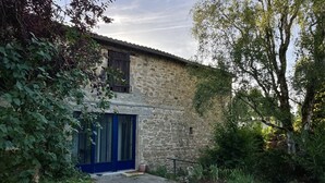 Edificio de piedra rústica con una puerta azul y una ventana pequeña, rodeado de follaje y un camino de grava.