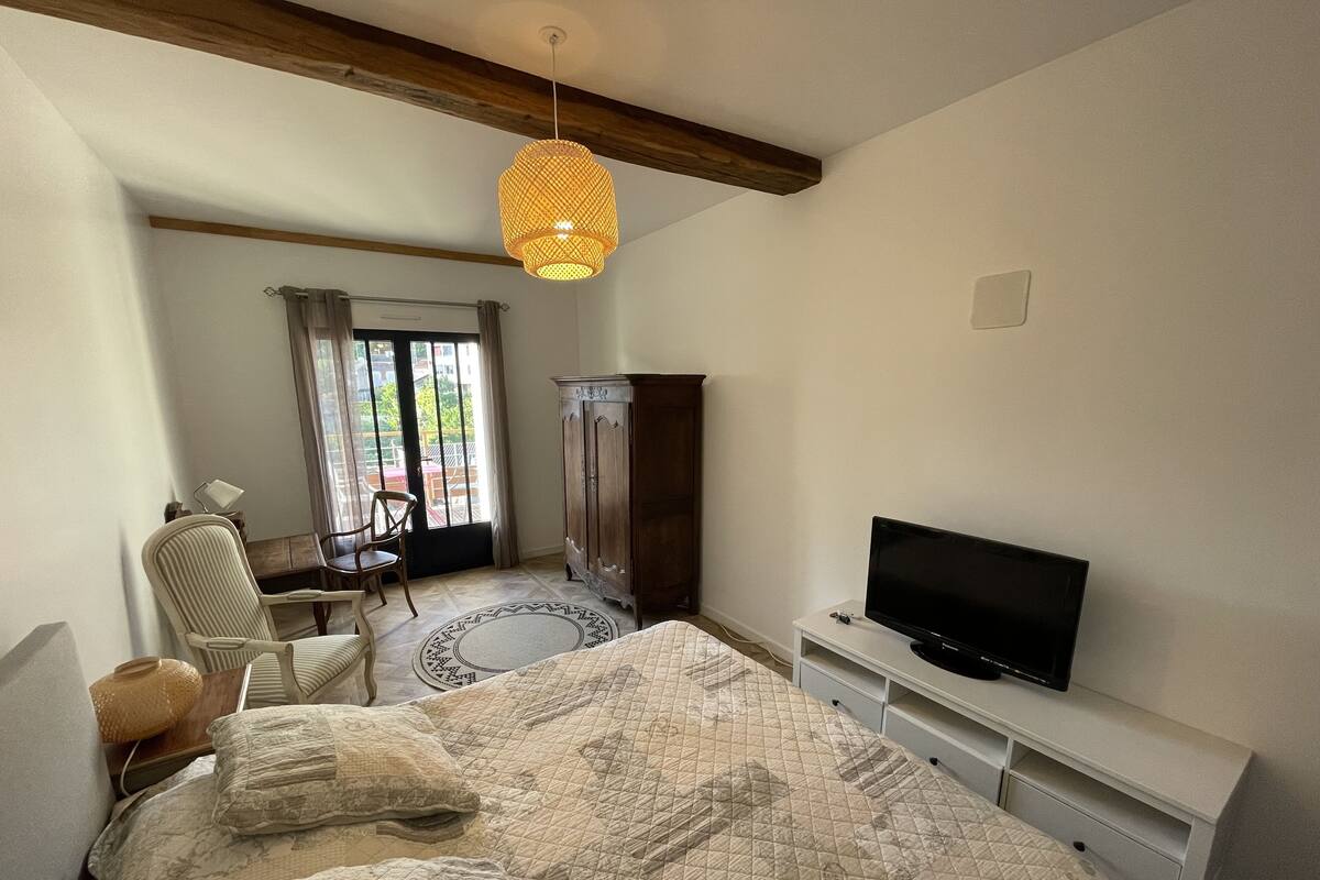 Quarto com uma cama grande, televisão em um suporte branco, guarda-roupa de madeira e uma cadeira perto da janela.