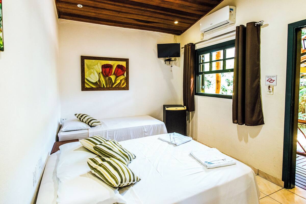 Un dormitorio con dos camas, un cuadro de flores, un equipo de aire acondicionado, una ventana con cortinas y una puerta.