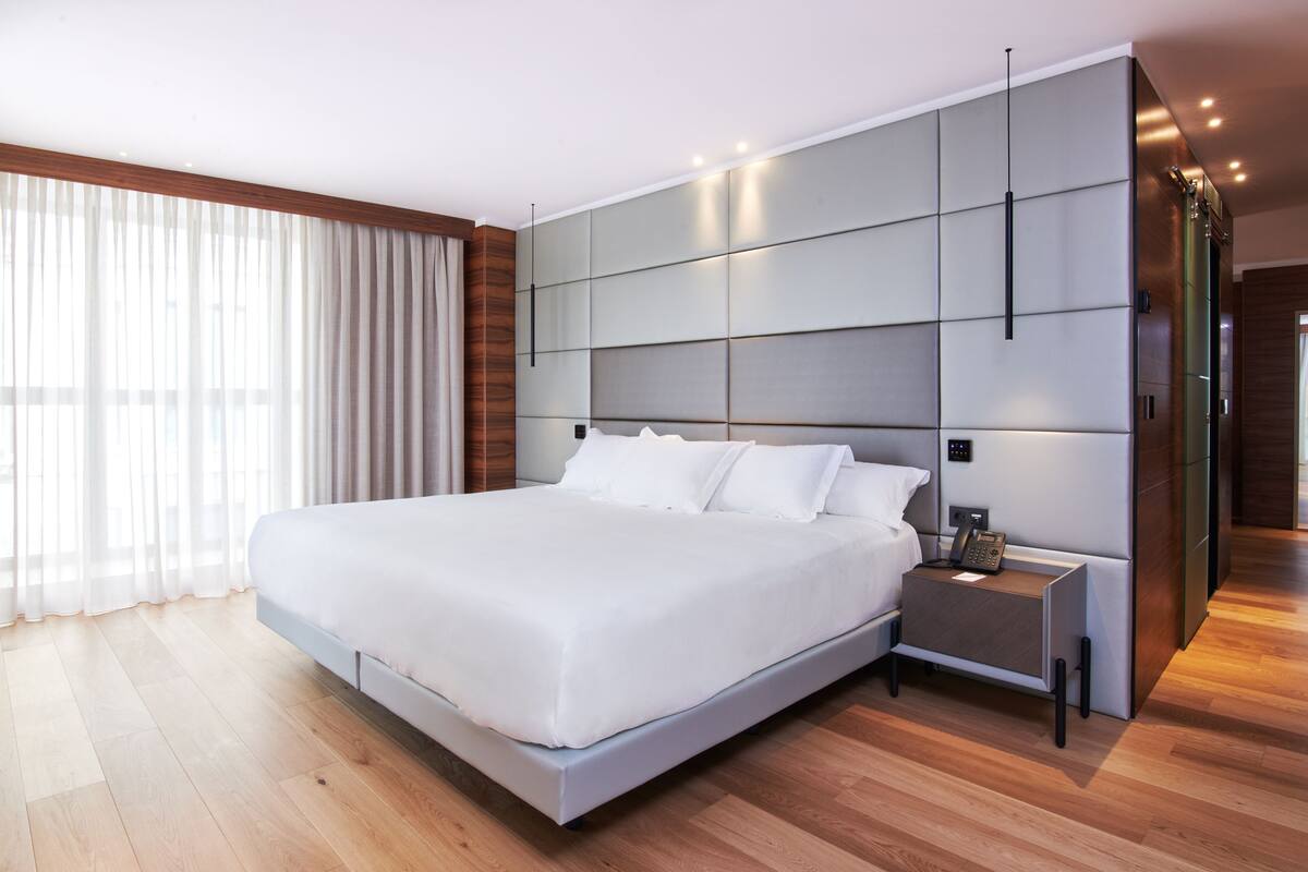 A modern bedroom with a large bed, a bedside table with a lamp, and a large window with curtains.