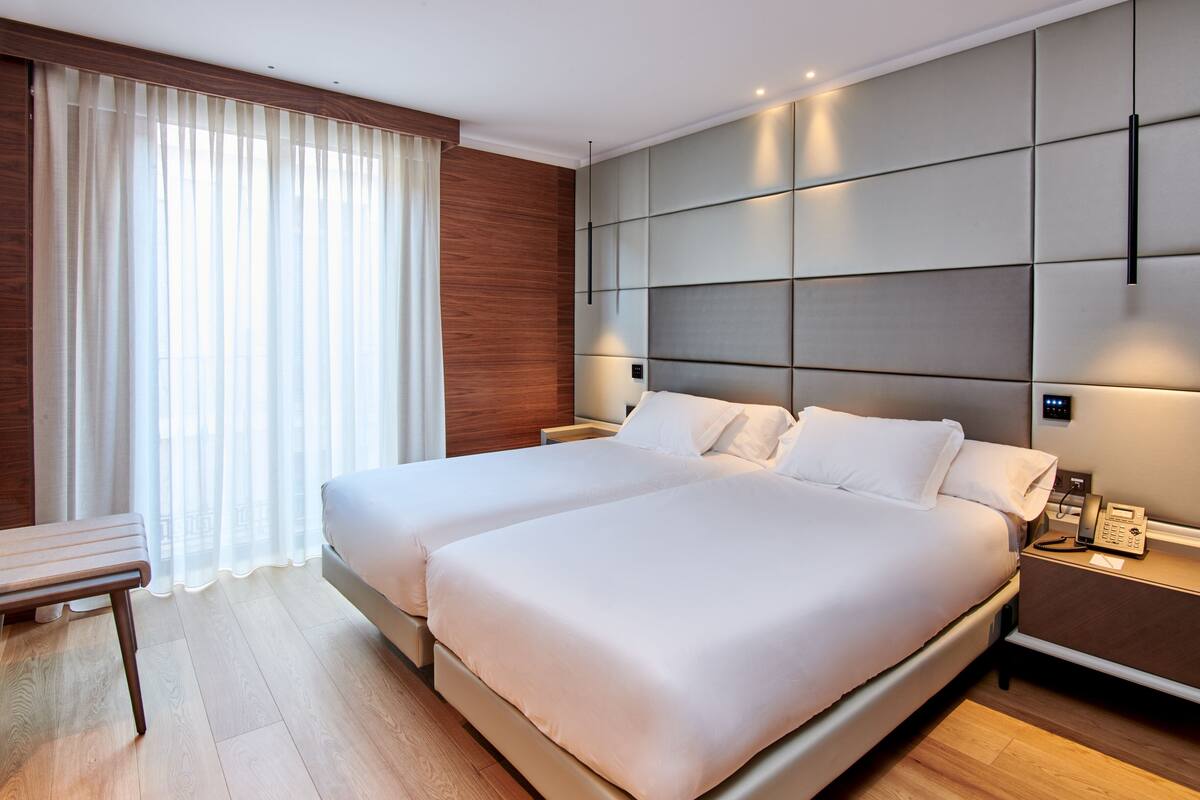 A modern hotel room with two beds, a bedside table, a chair, and a large window with curtains.