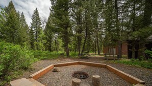 Un foyer circulaire, entouré de bancs en bois et bordé d’une zone de gravier, est aménagé dans un environnement boisé, avec de grands arbres et une structure en bois en arrière-plan.
