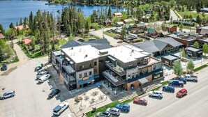 A multi-story building with a flat roof, a parking lot with several cars, and a view of a lake and forested hills in the background. - Downtown condo short walk to lake, patio and awesome common area (Grand Lake)